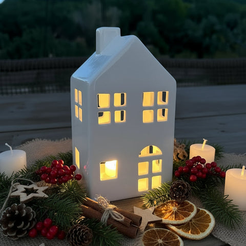 Decorative house-shaped lantern with lights, surrounded by candles and Christmas decorations on a table outdoors.