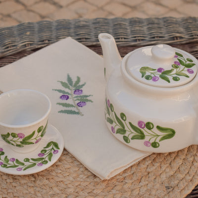Tea set with teapot, cup, and saucer featuring floral designs on a woven surface.