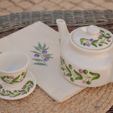 Tea set with teapot, cup, and saucer featuring floral designs on a woven surface.