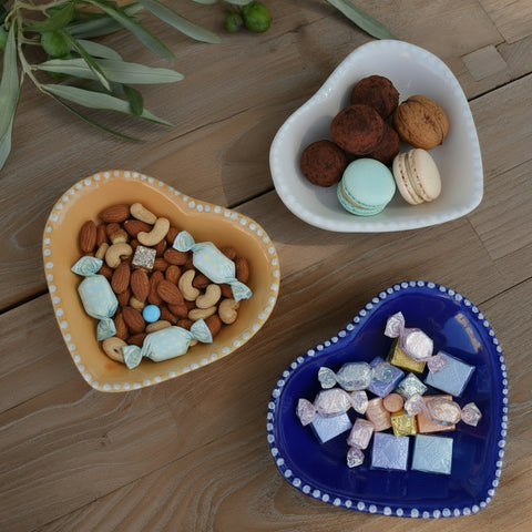 Three ceramic heart-shaped dishes in orange, white, and blue on a wooden surface with olive branches.