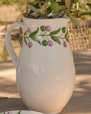 White pitcher with olive branch design on a wooden surface with olives and leaves.